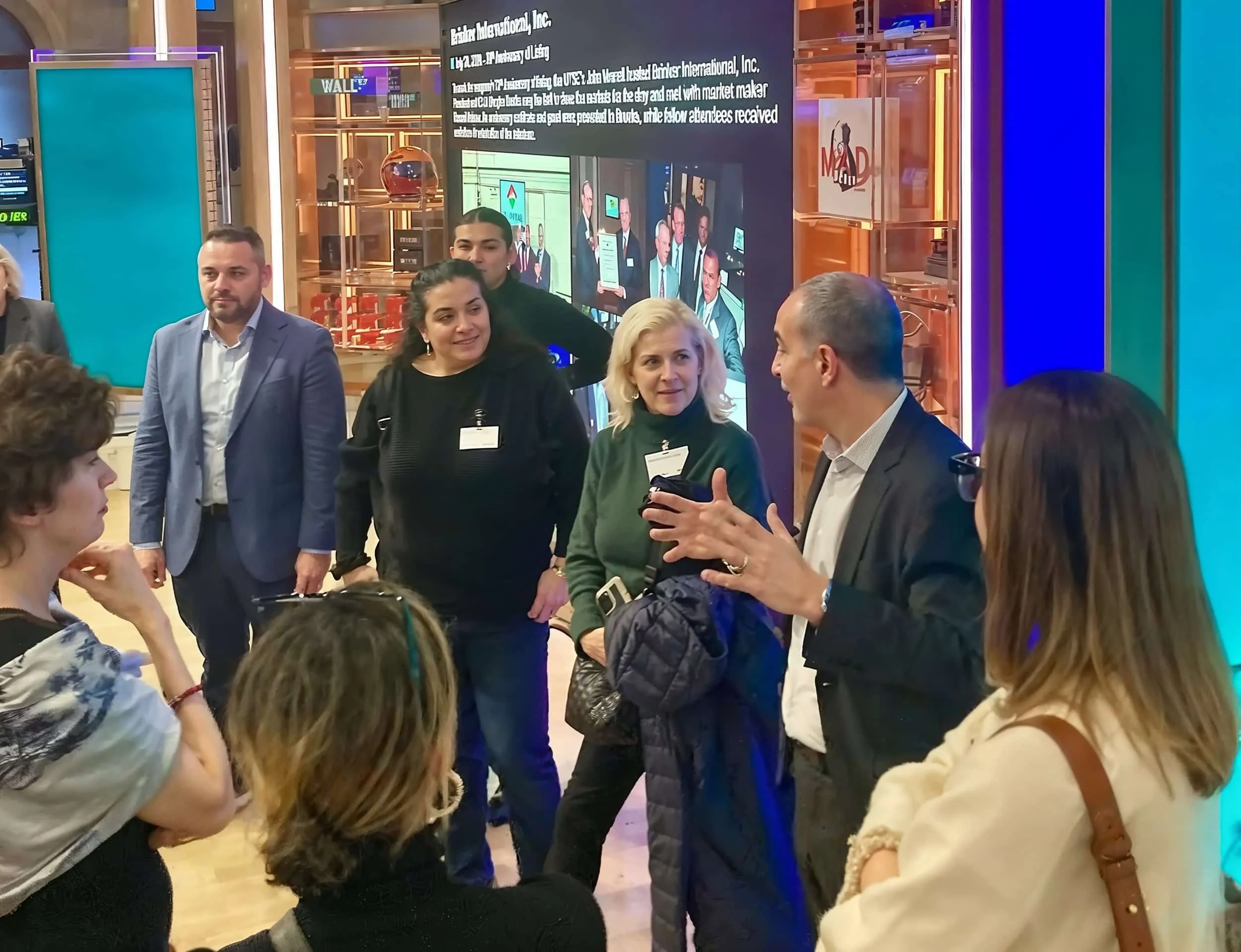 A group of diverse people engaging in a conversation at a professional event or conference, standing in a brightly lit, modern indoor setting with digital screens and displays in the background.