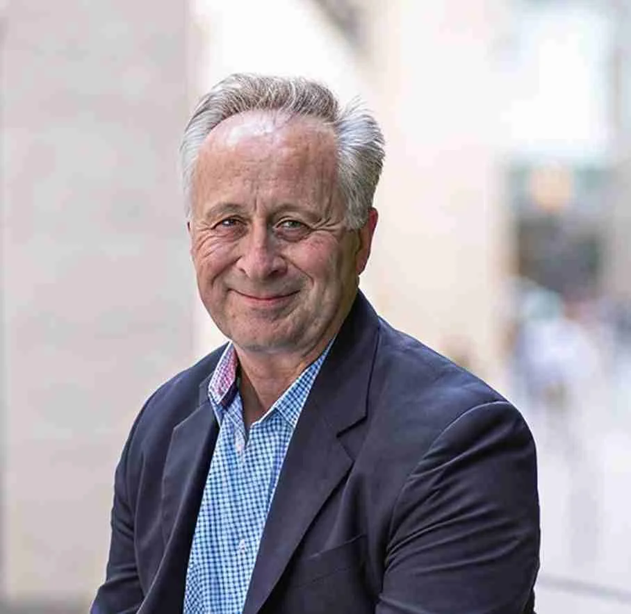 A smiling older man with gray hair, wearing a dark blazer and a checkered shirt, standing outdoors in an urban setting.