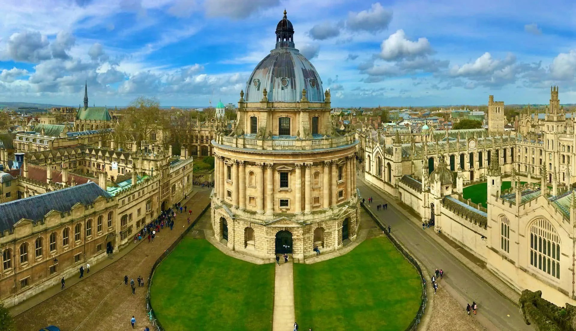 Aerial view of the Radcliffe Camera in Oxford, England, featuring a circular stone building with a domed roof, surrounded by historic university architecture and a green lawn in the foreground with people walking around.