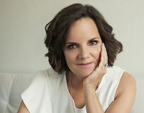 Woman with short, wavy brown hair wearing a white sleeveless top, resting her face on her hand, looking directly at the camera.