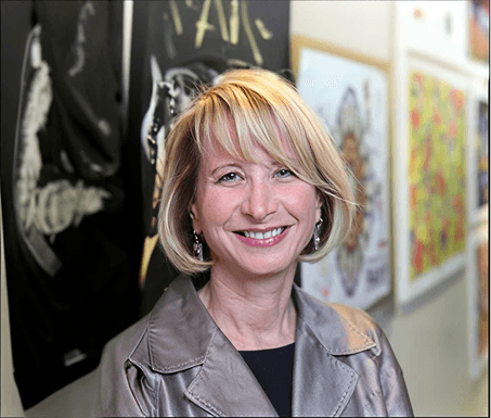 A middle-aged woman with blonde hair and a wide smile, wearing a gray jacket, standing in front of colorful artwork on a wall.