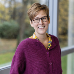 A woman with short hair and glasses smiling, wearing a purple cardigan and a colorful blouse, standing outdoors near a window with trees in the background.