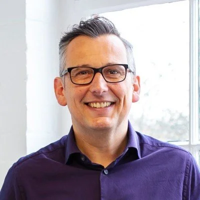 A smiling man with gray hair, glasses, and wearing a purple shirt, standing in front of a window with natural light.