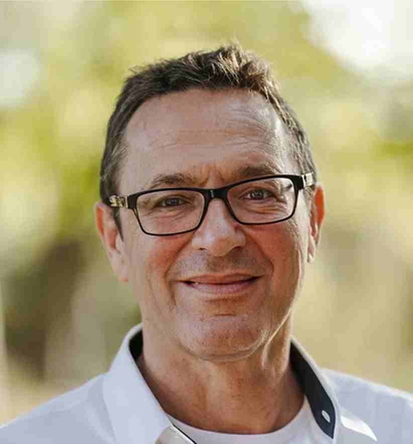 A middle-aged man with short brown hair, wearing glasses and a white shirt, smiling outdoors with a blurred green background