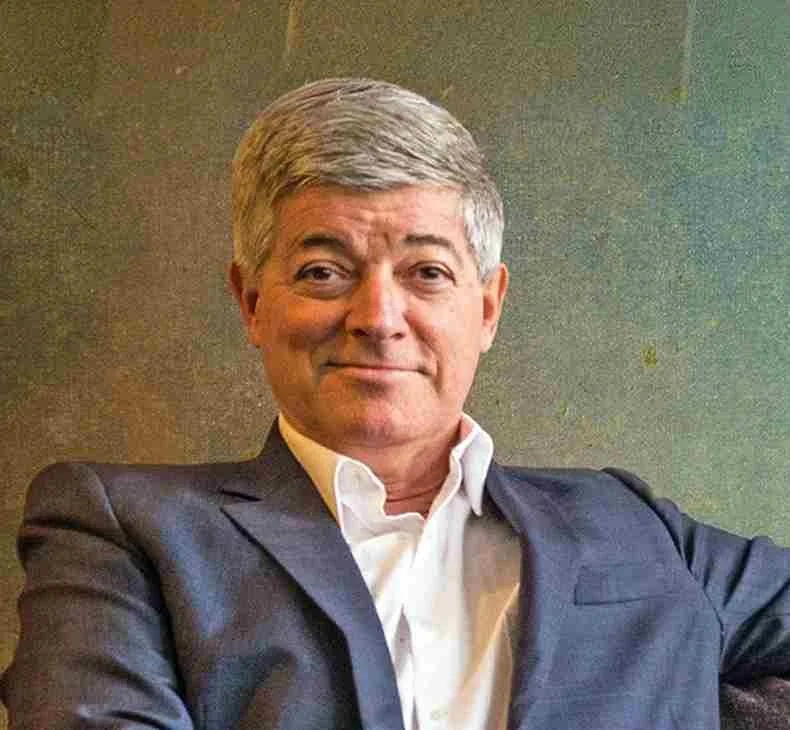 Portrait of an older man with gray hair, wearing a dark suit and white shirt, sitting and smiling at the camera.