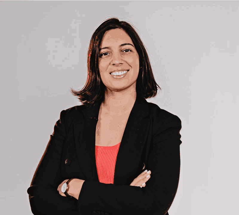A woman with shoulder-length brown hair, wearing a black blazer over a red top, smiling with arms crossed against a light gray background.