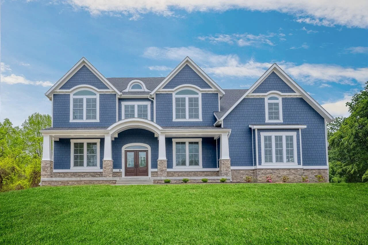 A large two-story blue house with white trim and a stone foundation, set on a green lawn with trees in the background and a blue sky with some clouds above.