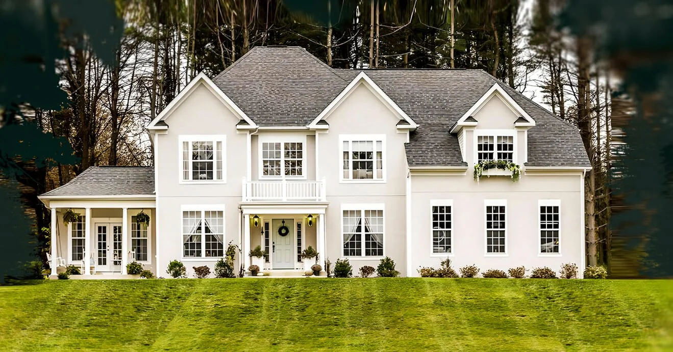 Large white two-story house with multiple windows, a front porch with chairs, and a well-maintained lawn, set against a background of trees.