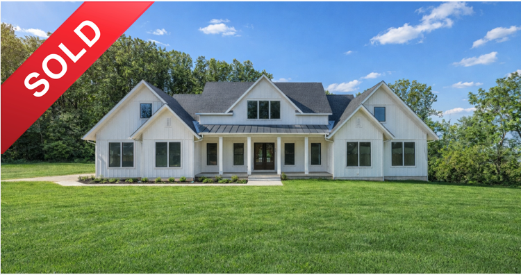 Large white house with multiple gables and a front porch, sold sign in the top left corner, green lawn, blue sky with some clouds, surrounded by trees.