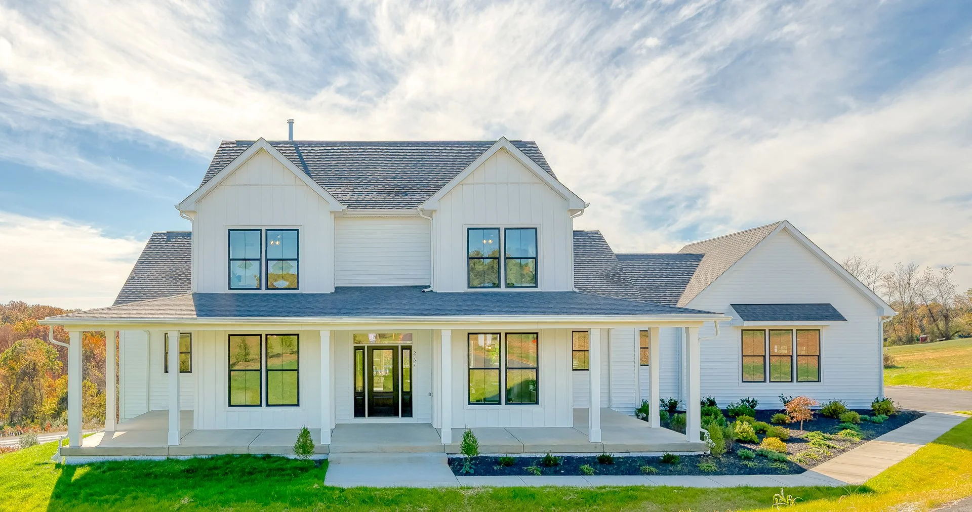 A two-story white house with a front porch, multiple windows, and a gabled roof, surrounded by a landscaped yard with green grass and plants.