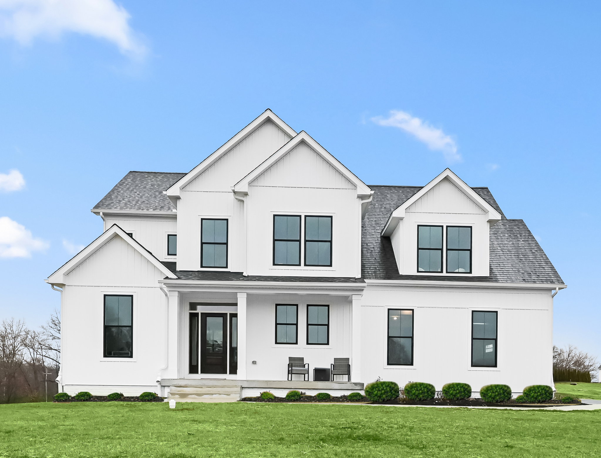 White modern two-story house with black window frames, front porch with two chairs, surrounded by green grass and bushes, under a blue sky with a few clouds.