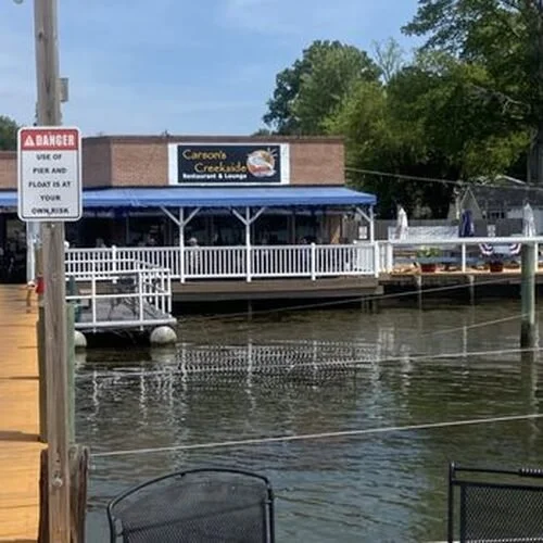 View of Carson's Creekside restaurant and lounge by the water with a dock, outdoor seating, and a sign warning about the danger and the use of pier and float.