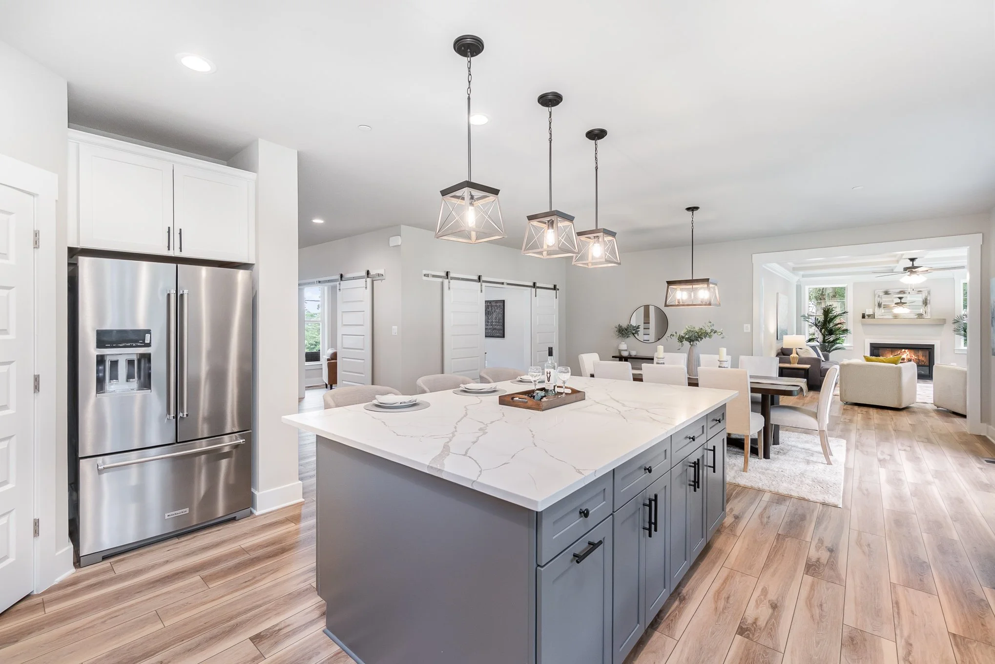 Open-concept kitchen and dining area with white cabinetry, gray kitchen island with marble countertop, modern pendant lighting, light wood flooring, and living room with fireplace and seating in the background.