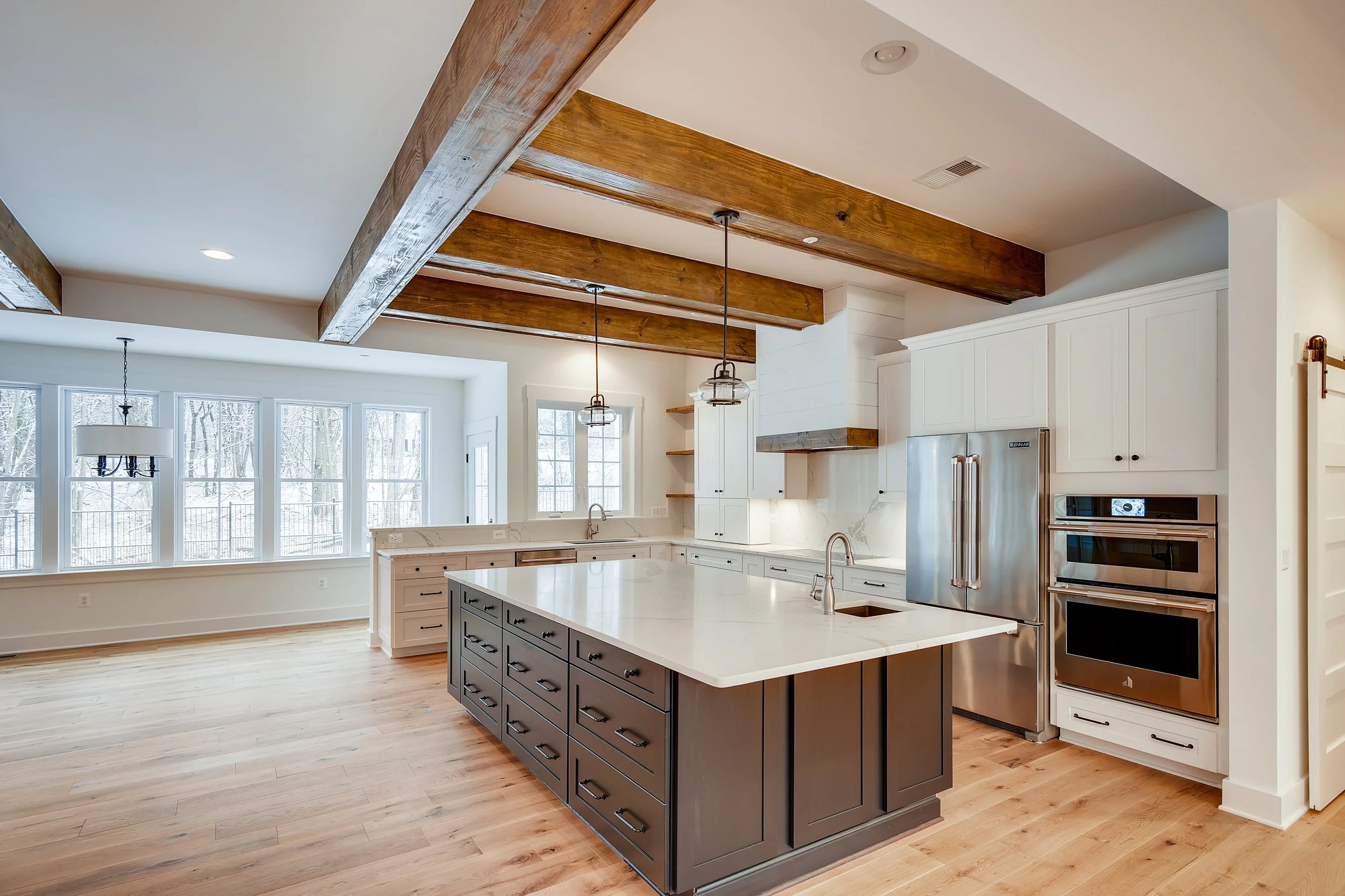 Modern kitchen with white cabinets, stainless steel appliances, dark kitchen island, wooden ceiling beams, and large windows with a snowy outdoor view.