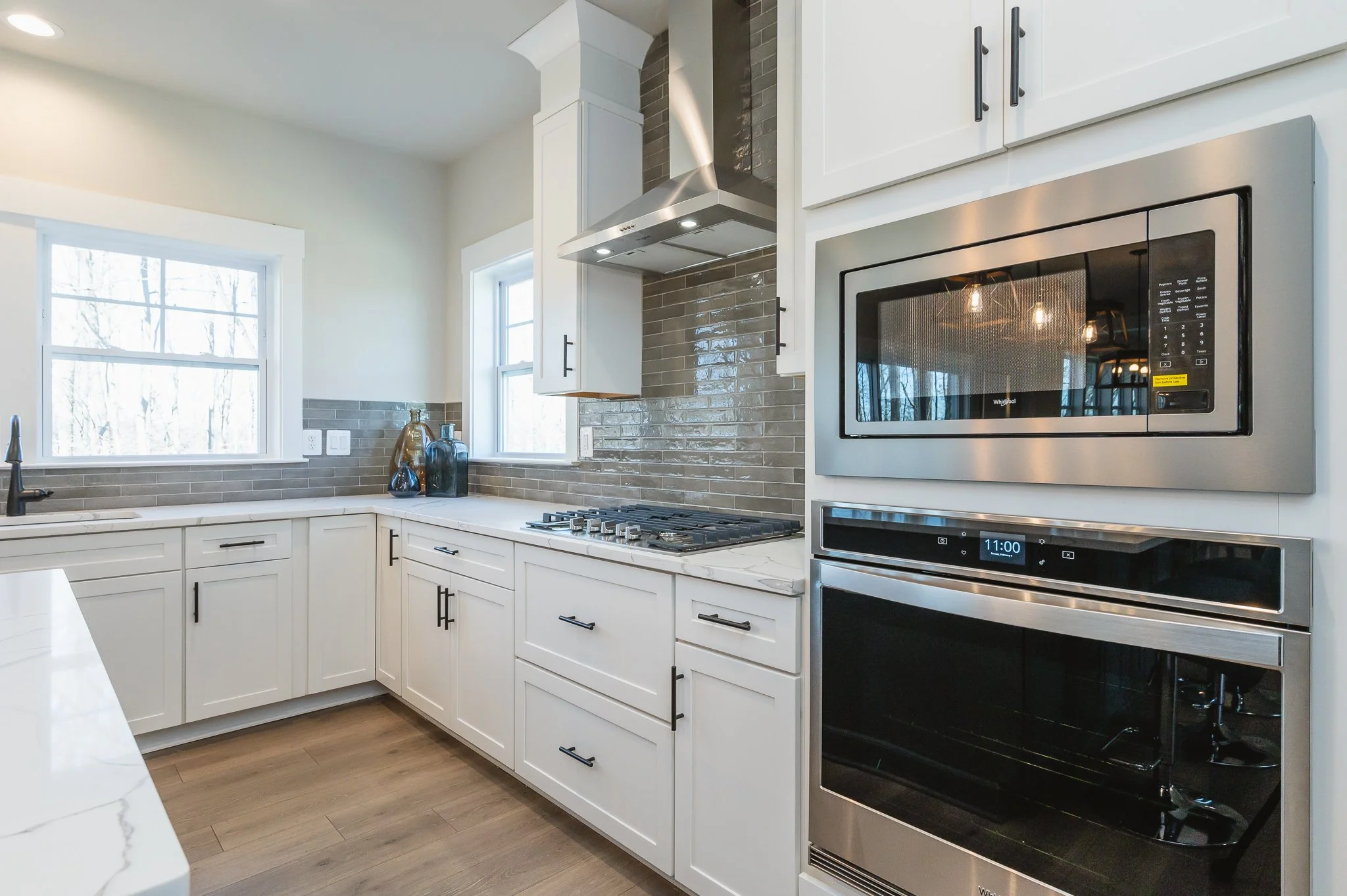Modern kitchen with white cabinets, a gas stove, stainless steel microwave and oven, a black sink, and wood flooring, with two windows providing natural light.