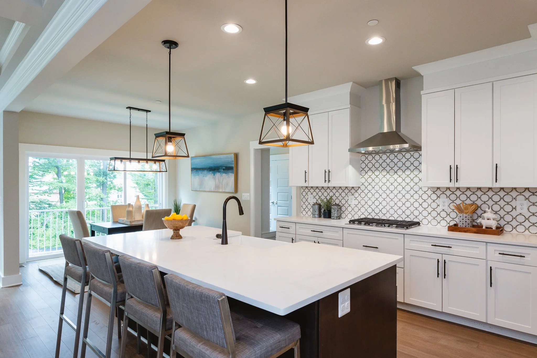 Modern kitchen with white cabinets, a large island with a white countertop, pendant lighting, and a dining area with sliding glass doors leading to a balcony with green trees outside.