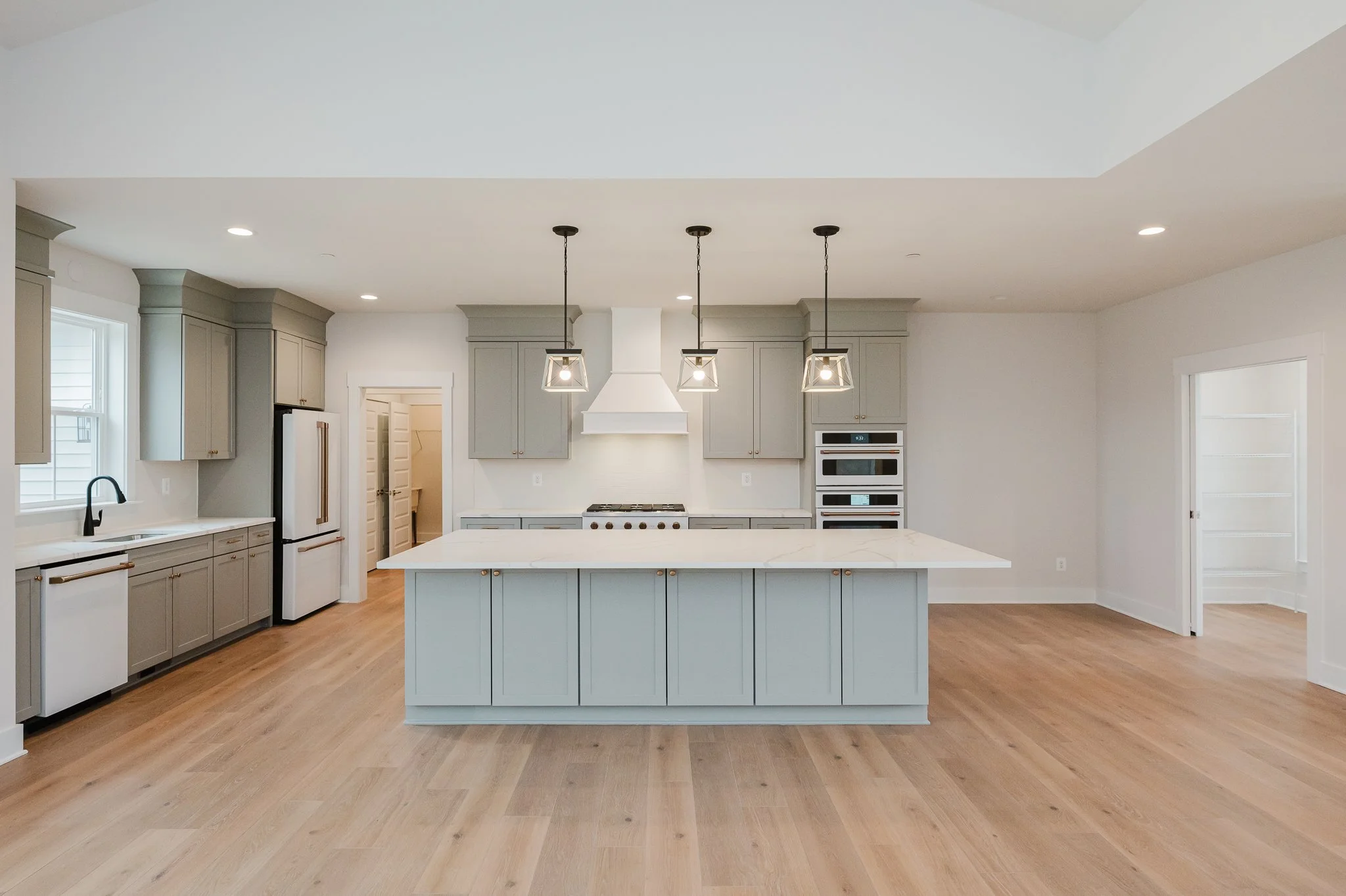 Empty modern kitchen with light gray cabinets, white marble island, hardwood floors, black pendant lights, and stainless steel appliances.