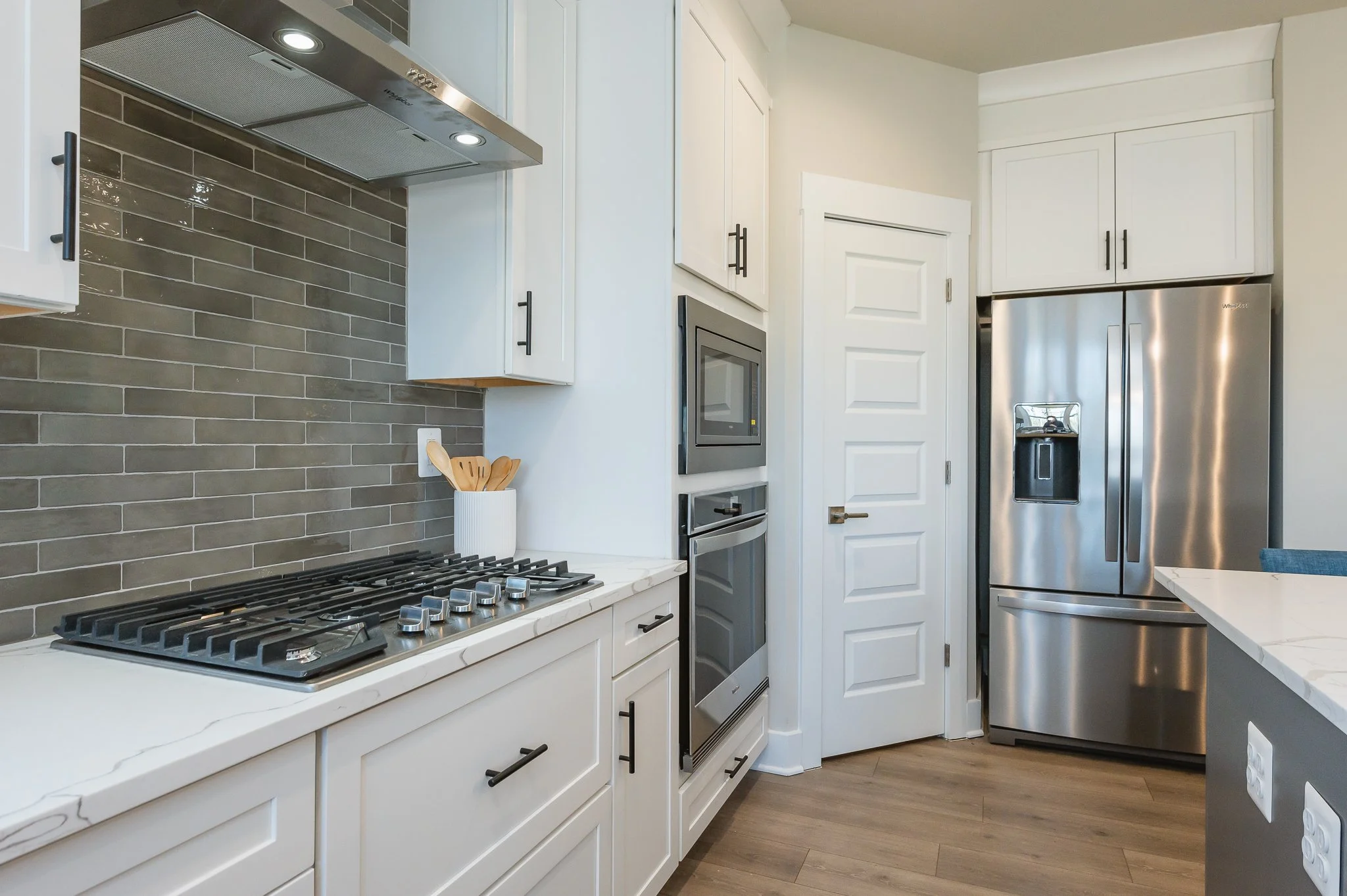 Modern kitchen with white cabinets, gray tile backsplash, stainless steel refrigerator, built-in microwave and oven, gas cooktop, and wood flooring.