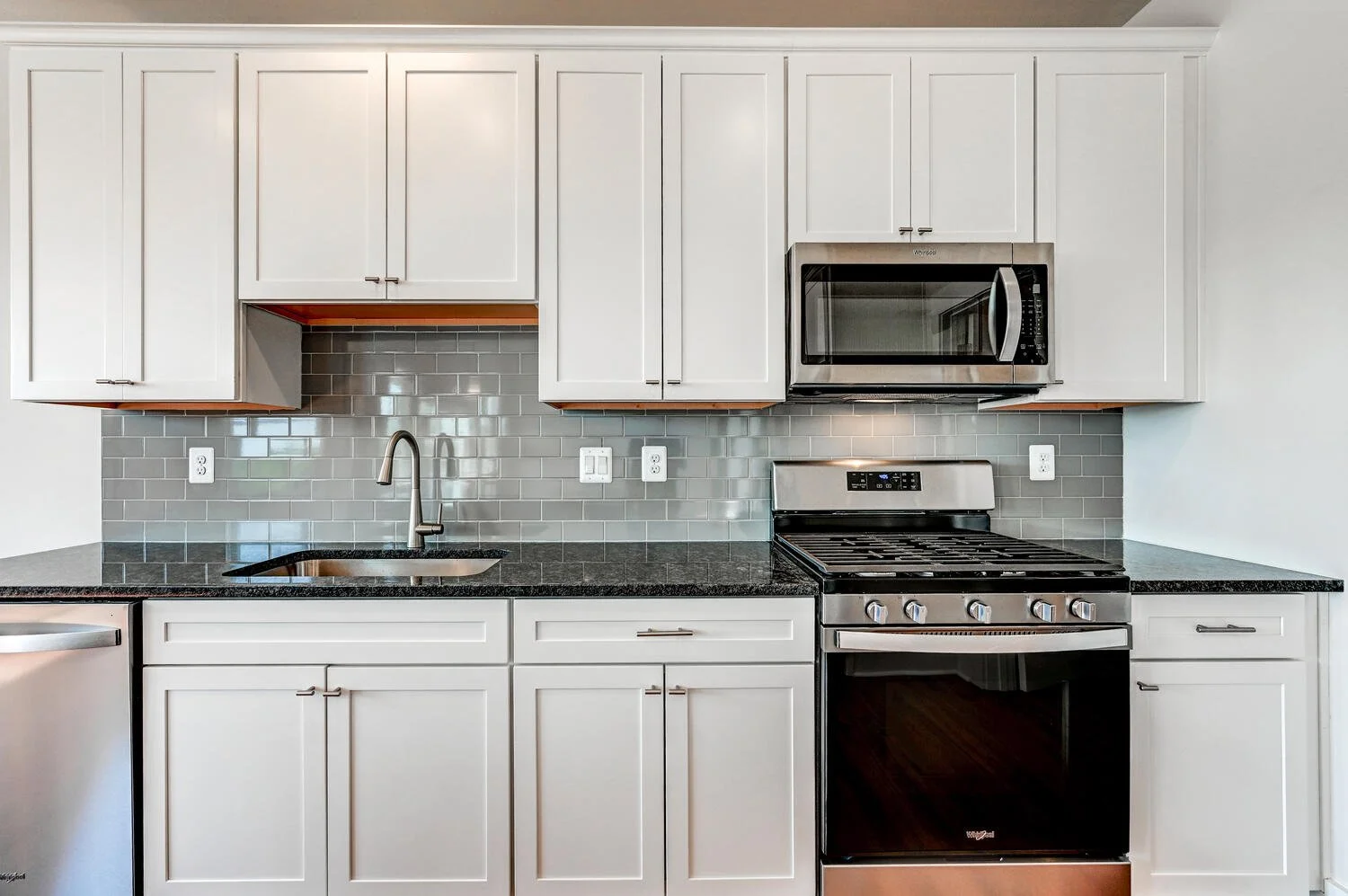 Modern kitchen with white cabinets, black granite countertops, stainless steel appliances, gray subway tile backsplash, and a sink with a tall faucet.