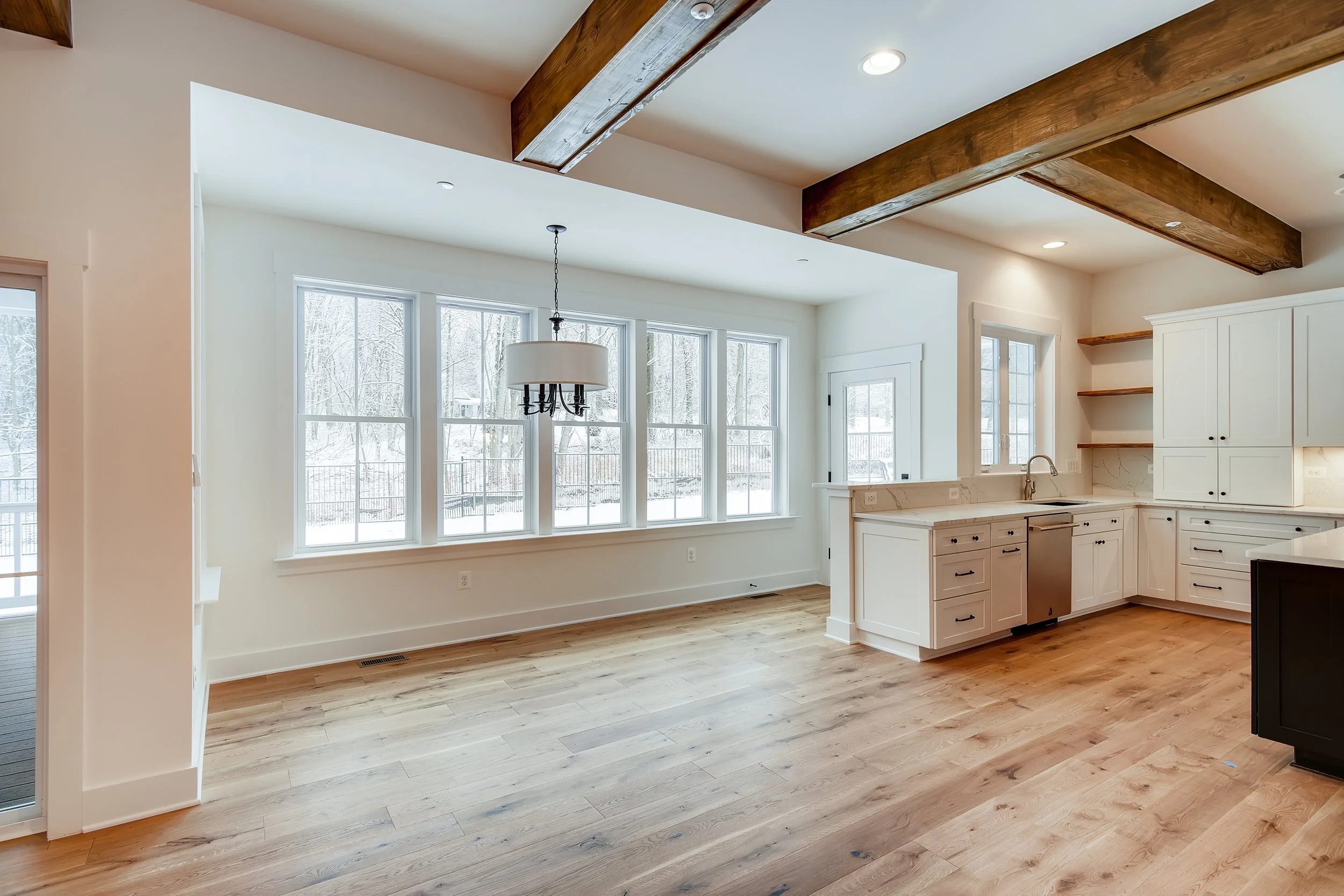 Empty kitchen and dining area with white cabinets, wooden beams, hardwood floors, and large windows showing a snowy outdoor scene.