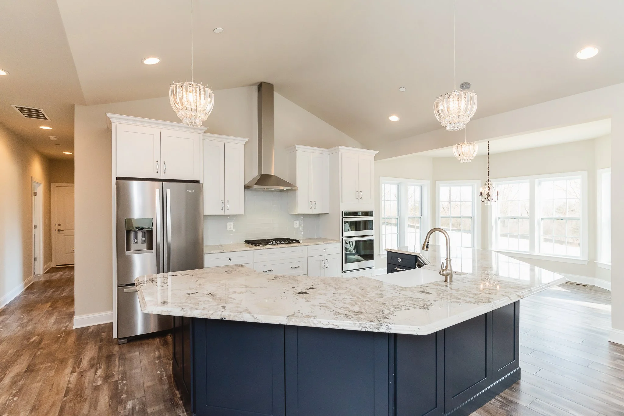 Modern kitchen with white cabinets, stainless steel refrigerator, granite island, and a breakfast area with large windows.