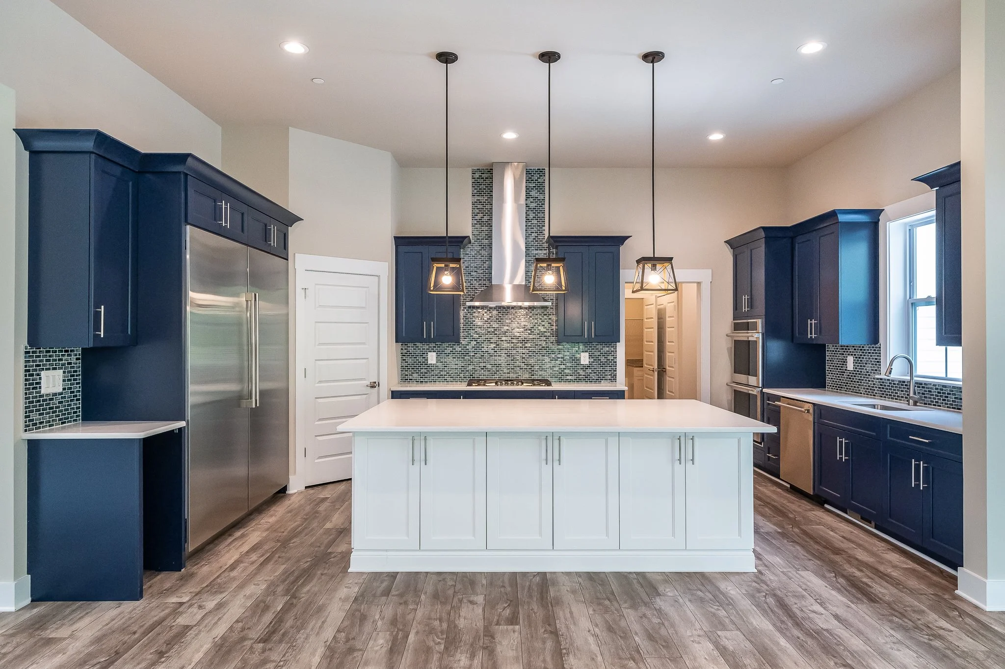 Modern kitchen with navy blue cabinets, a white kitchen island, stainless steel appliances, pendant lights, mosaic tile backsplash, and wooden flooring.
