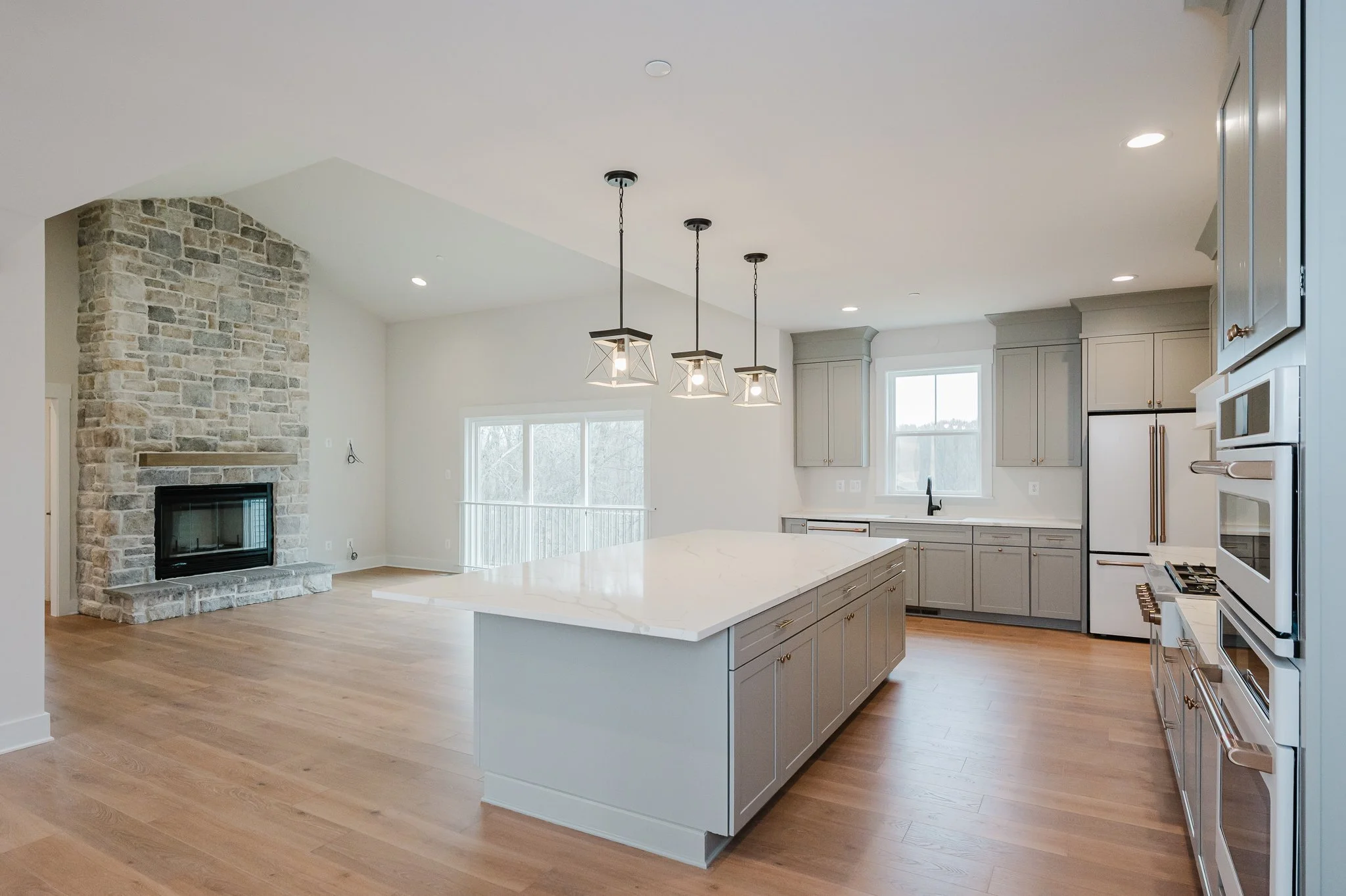Modern kitchen with light gray cabinets, a large white island with marble countertops, wooden flooring, and a stone fireplace in the open living area. Includes large windows and pendant lighting above the island.
