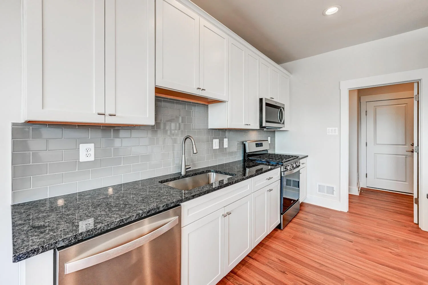 Modern kitchen with white cabinets, gray subway tile backsplash, black granite countertops, stainless steel appliances including a dishwasher, oven, and microwave, and hardwood floors.