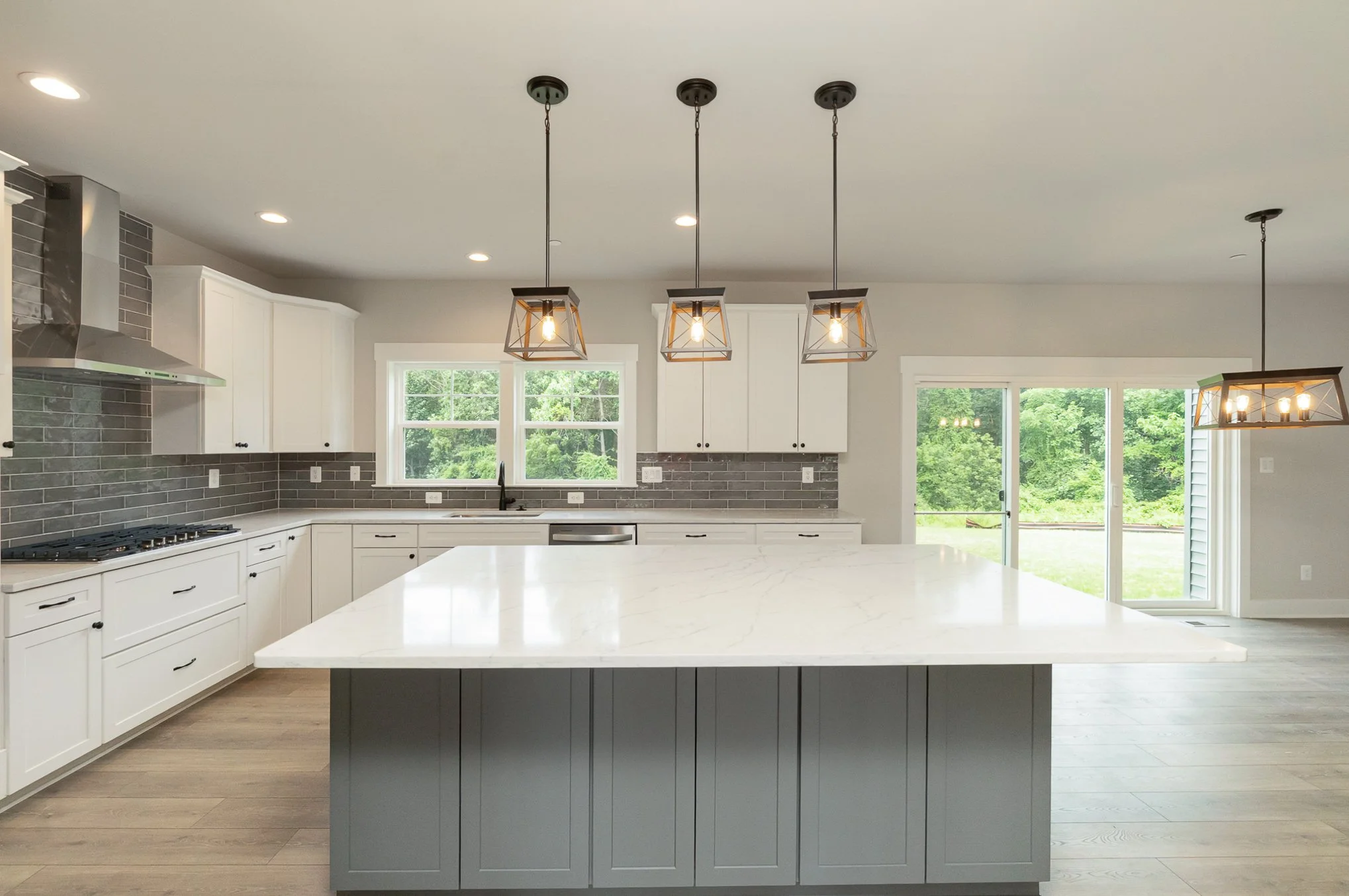 Modern kitchen with white cabinets, gray brick backsplash, large white island with gray base, three pendant lights above island, large window over sink, sliding glass door leading outside, light wood flooring.