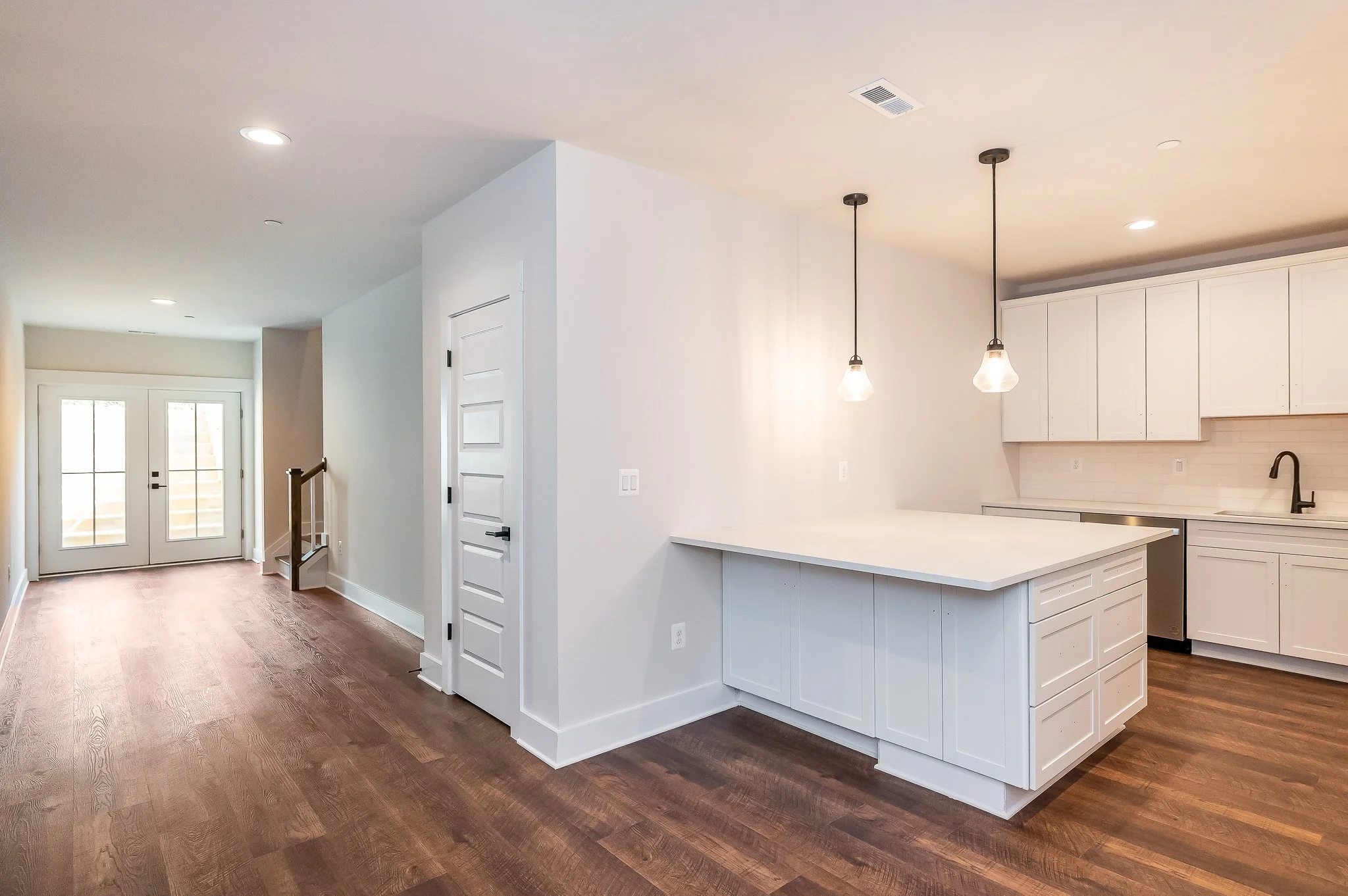 Empty modern kitchen with white cabinets, island, black faucet, and pendant lights, adjacent to a hallway with wooden floors and glass double doors.