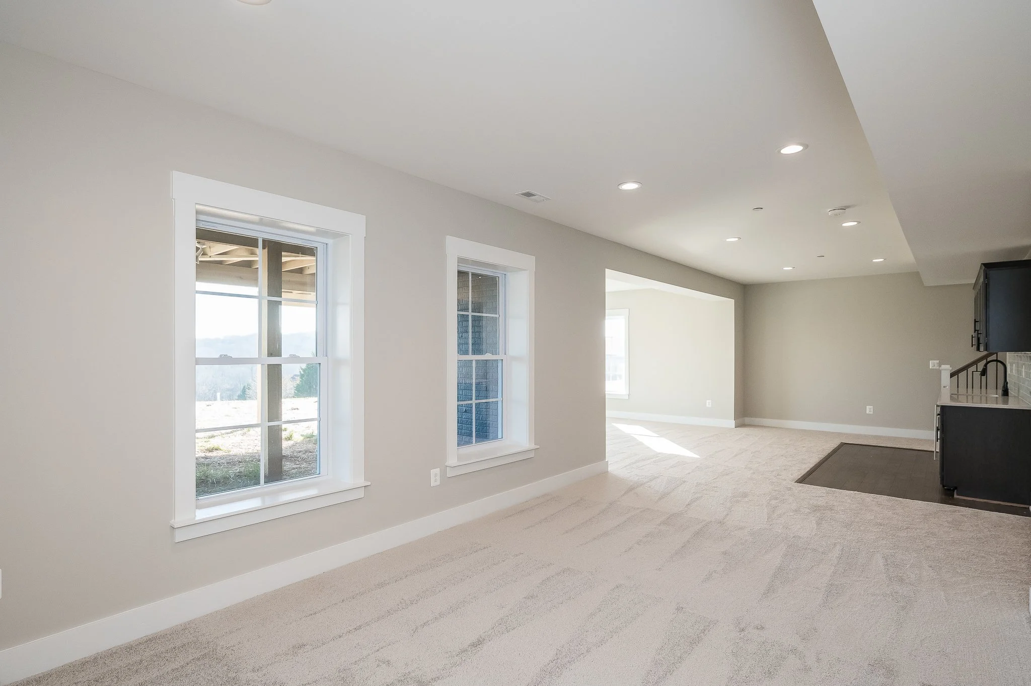Empty living room with beige walls, three windows, and cream carpet, sunlight coming through windows, visible part of kitchen to the right.