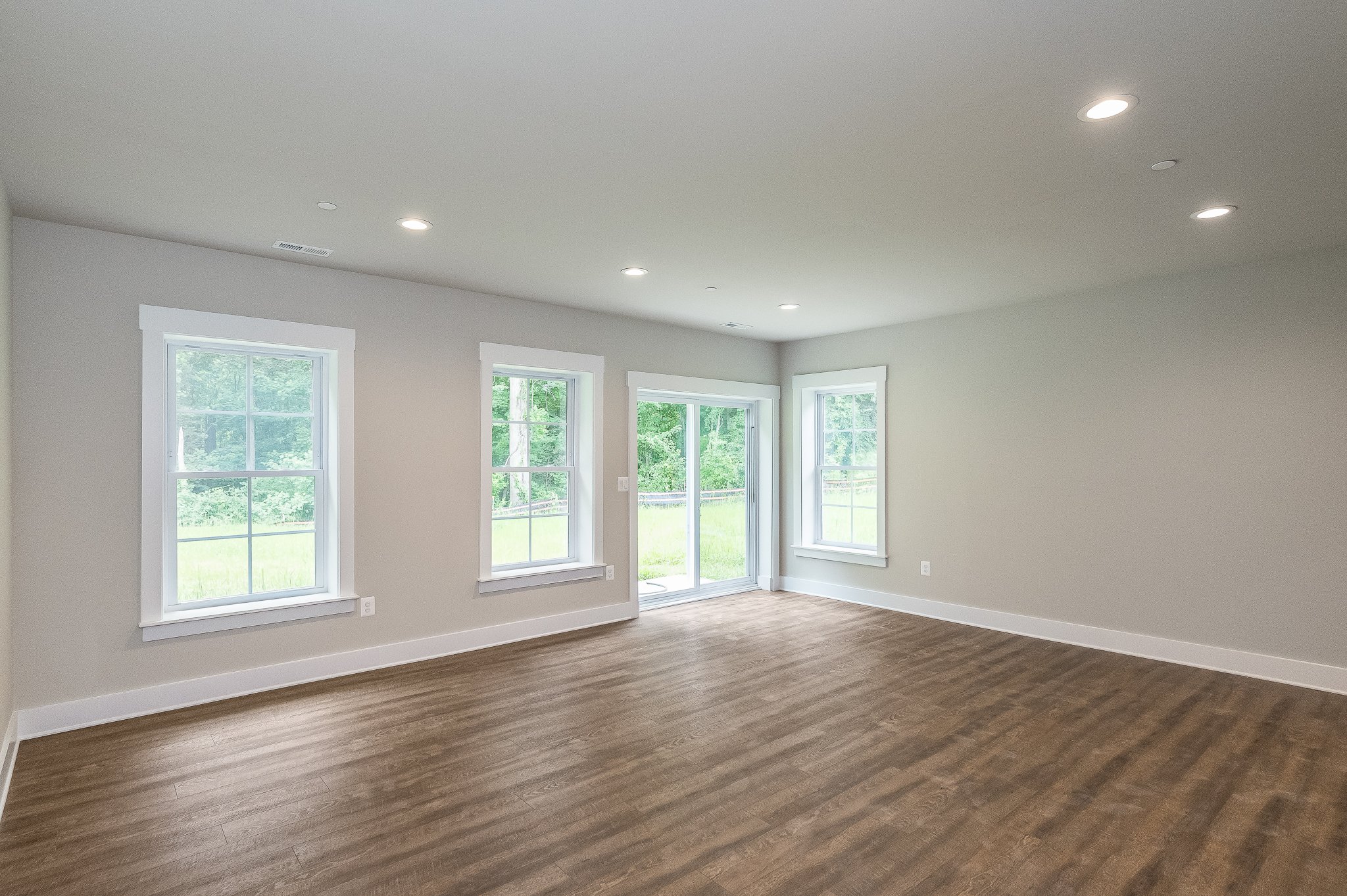 Empty living room with hardwood floors, four windows, white trim, and sliding glass door leading outside, with ceiling lights.