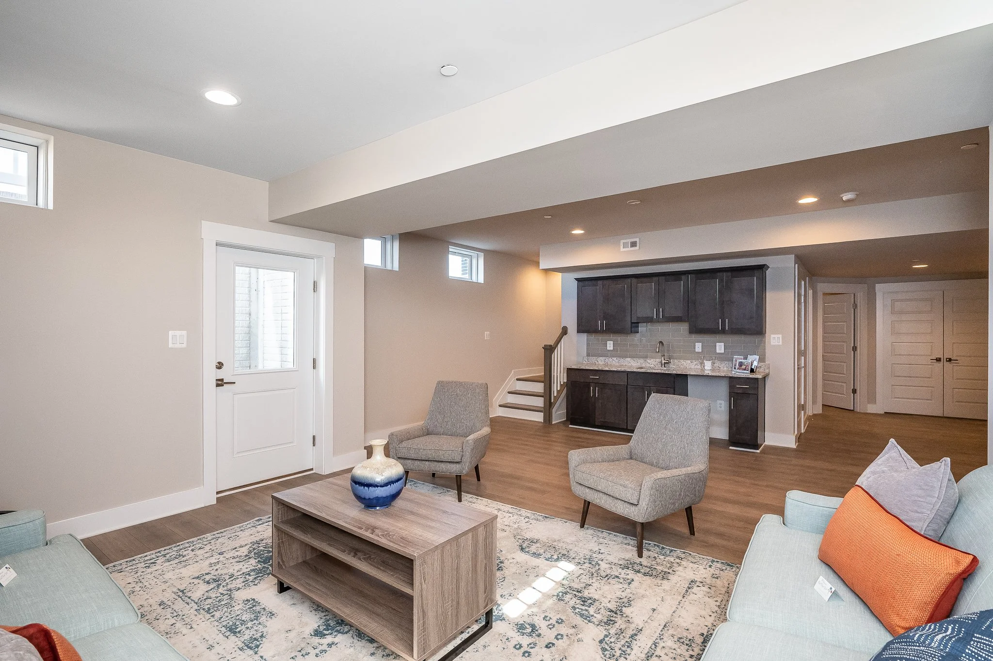 Modern living room with beige walls, hardwood floors, two gray armchairs, a light blue sofa with pillows, a wooden coffee table, and a kitchen area with dark cabinets and a granite counter.