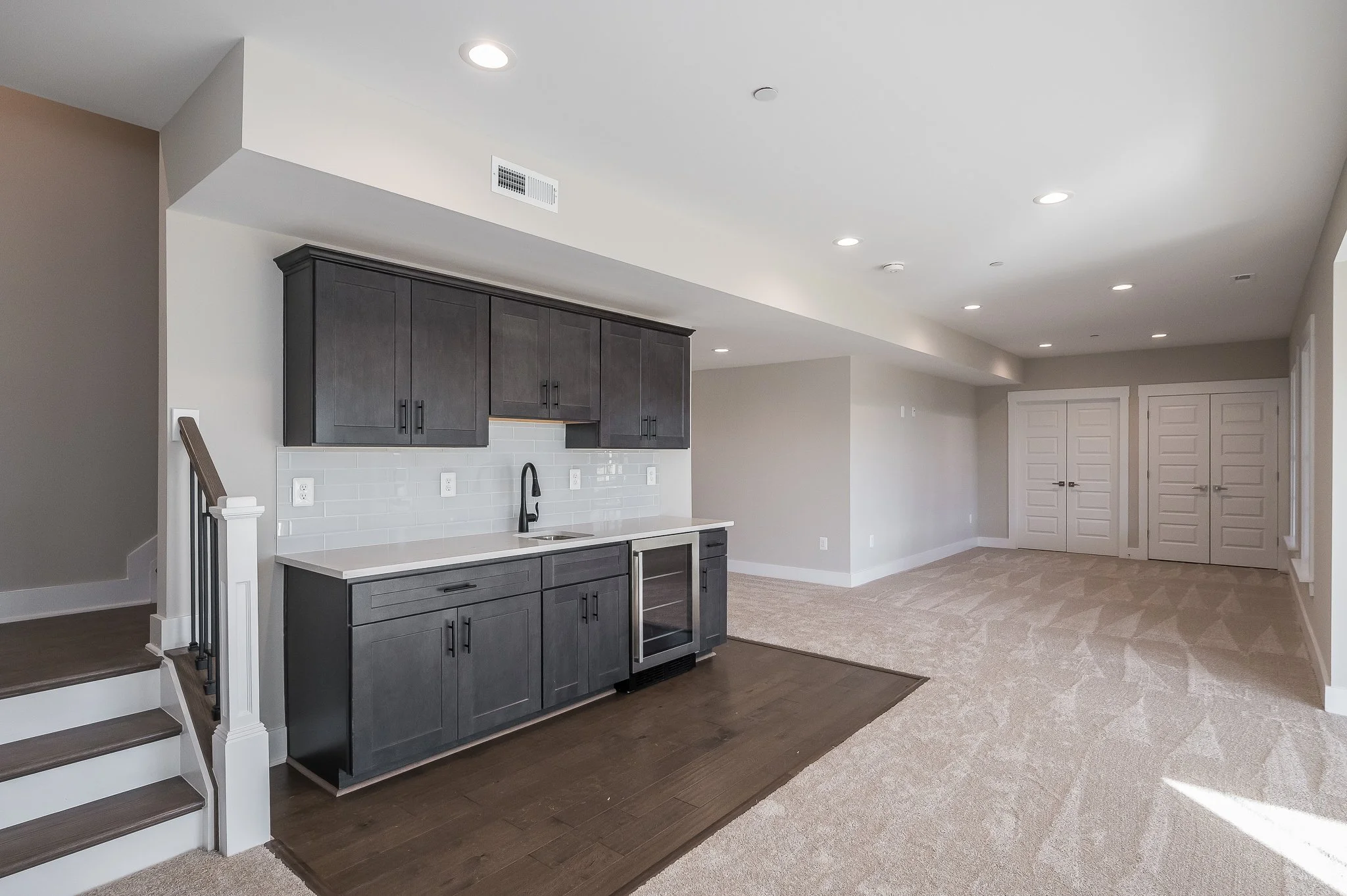 Empty living space with a kitchenette featuring dark cabinetry, a small refrigerator, white countertop, and a black faucet, adjacent to beige carpeted floor and a staircase on the left.