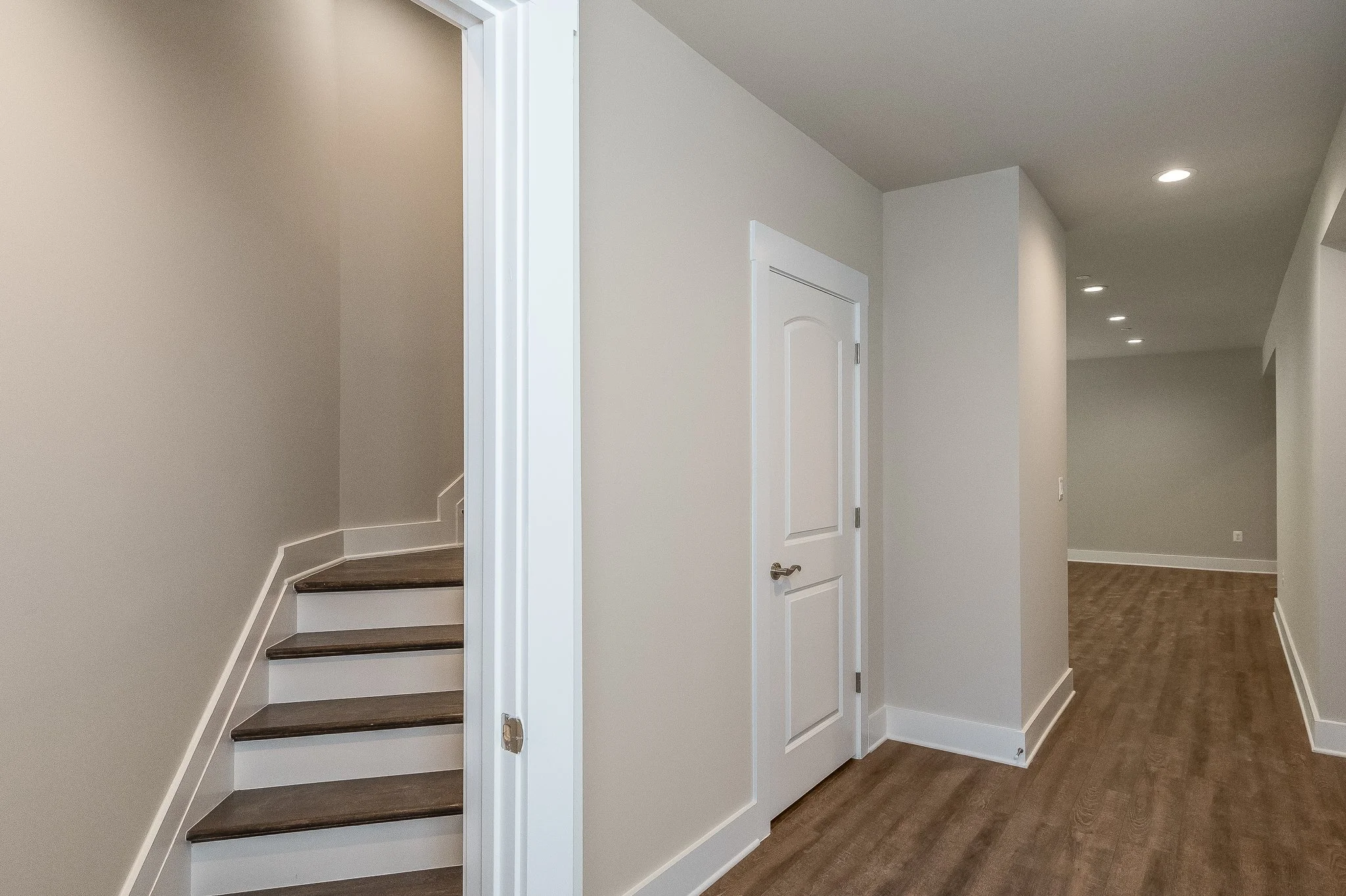 Interior of a modern home with a staircase, white door, hardwood floors, and recessed ceiling lights.