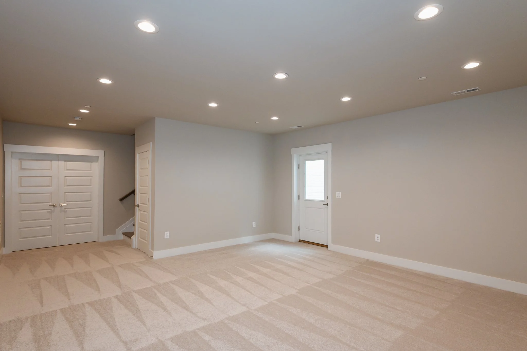 Empty living room with beige carpet, white walls, and a door with a window, illuminated by recessed ceiling lights.