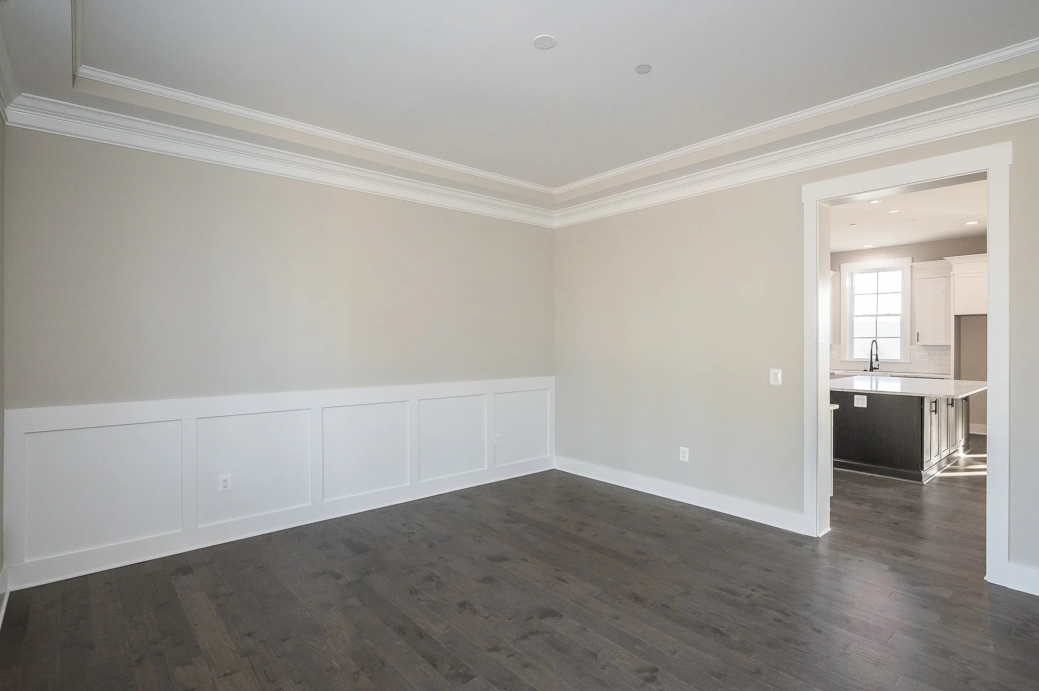 Empty living room with beige walls, dark hardwood flooring, crown molding, baseboards, and a view into the kitchen with a window and island.