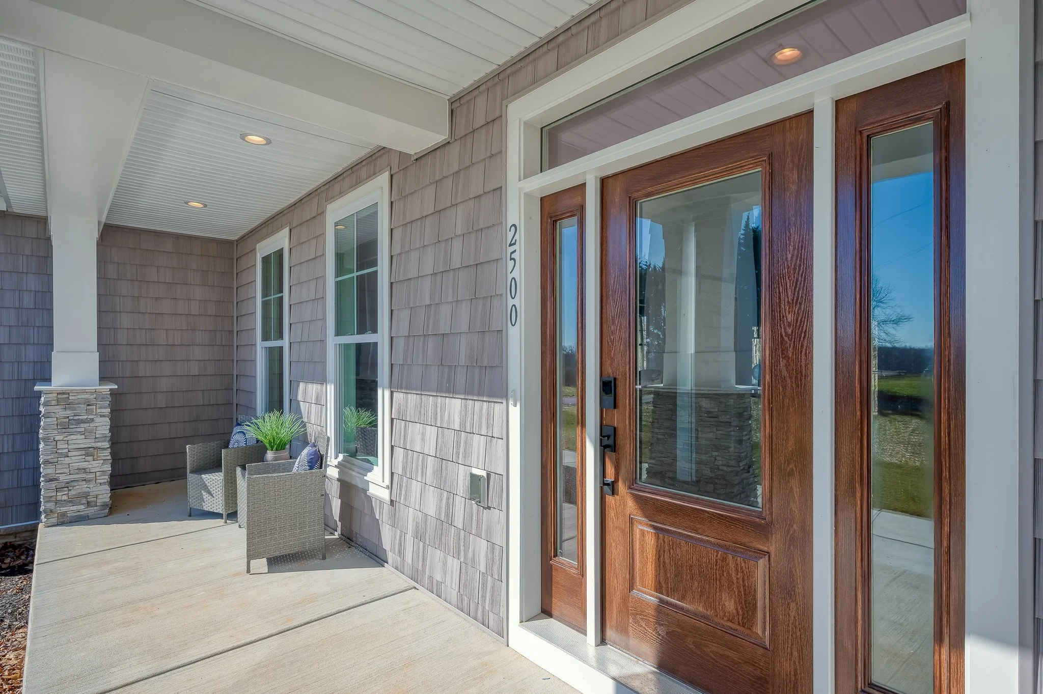 Front porch with a wooden door, two windows, and two chairs with pillows and potted plants.