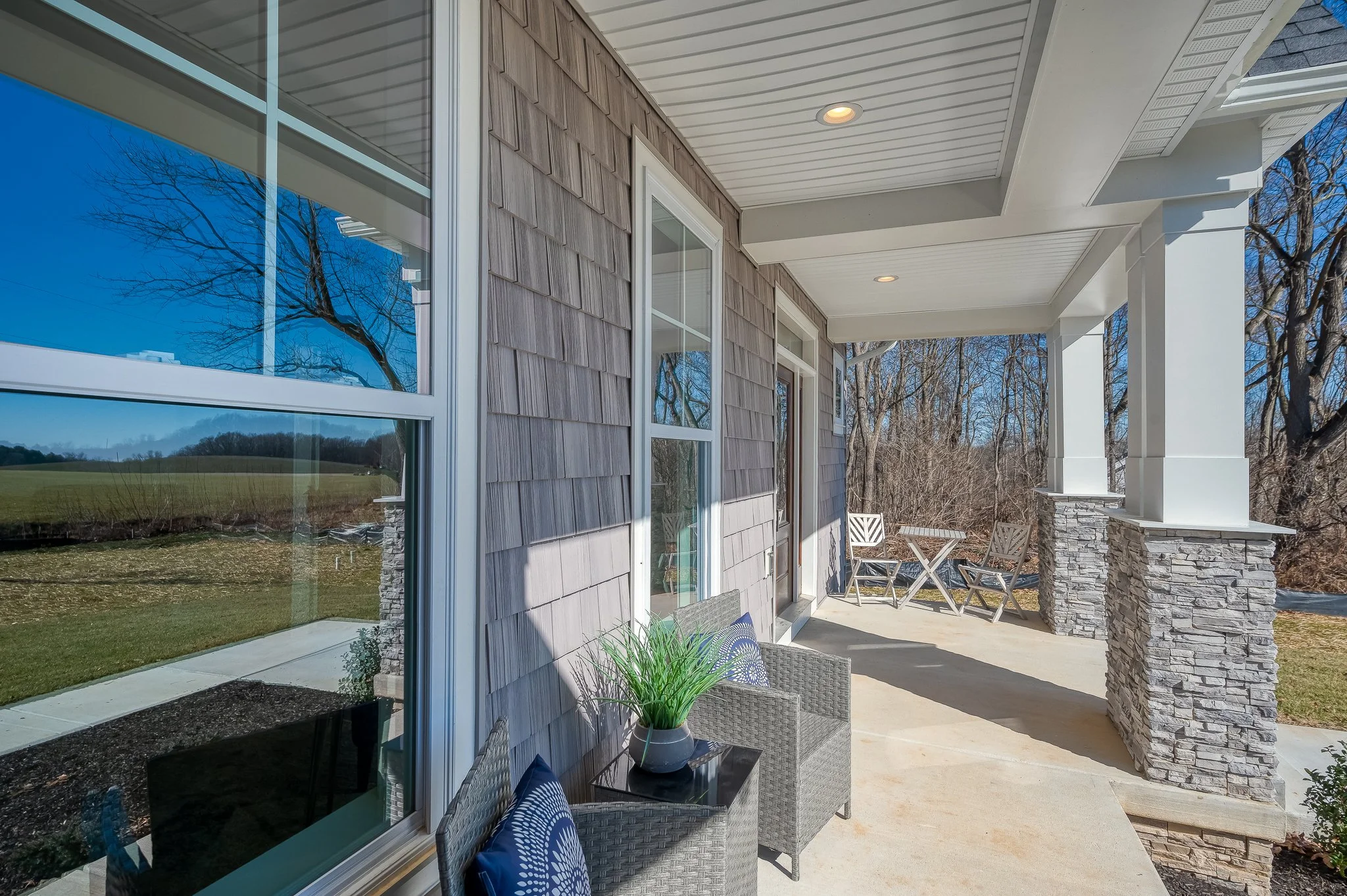 View of a house's front porch with chairs, table, and decorative stone pillars, overlooking a yard with trees and open fields on a sunny day.