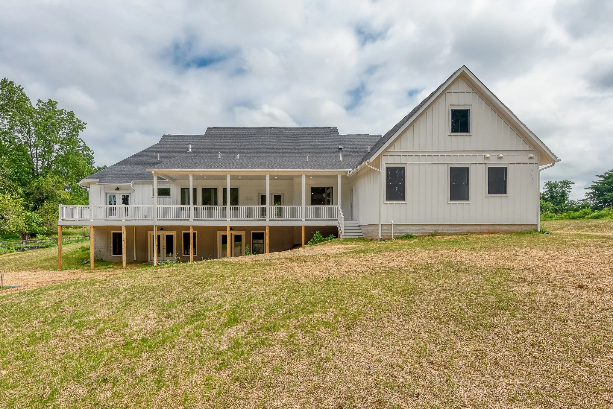 View of a modern white house with a spacious deck, multiple windows, and an unfinished yard under a cloudy sky.
