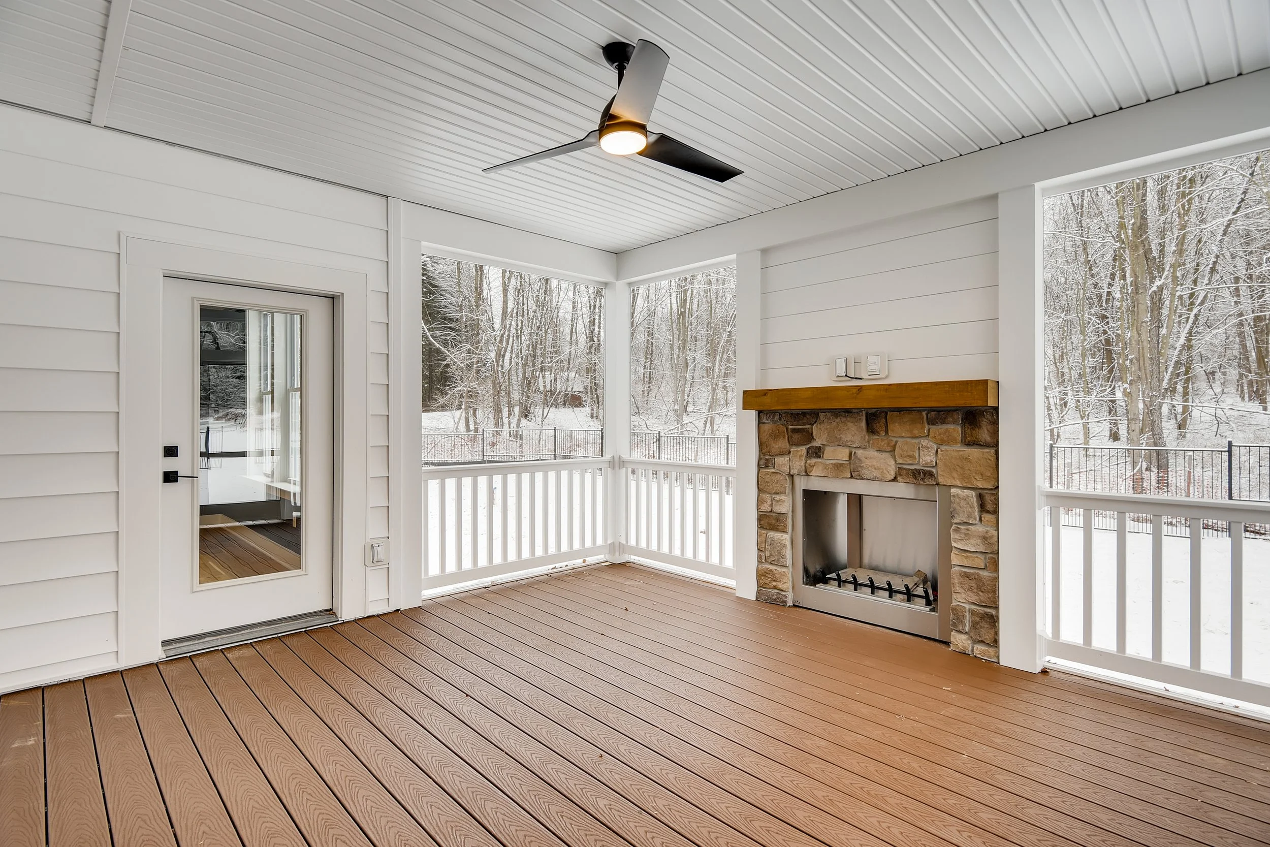 Empty enclosed porch with wooden floor, white siding, and a stone fireplace, overlooking a snowy outdoor scene with trees.