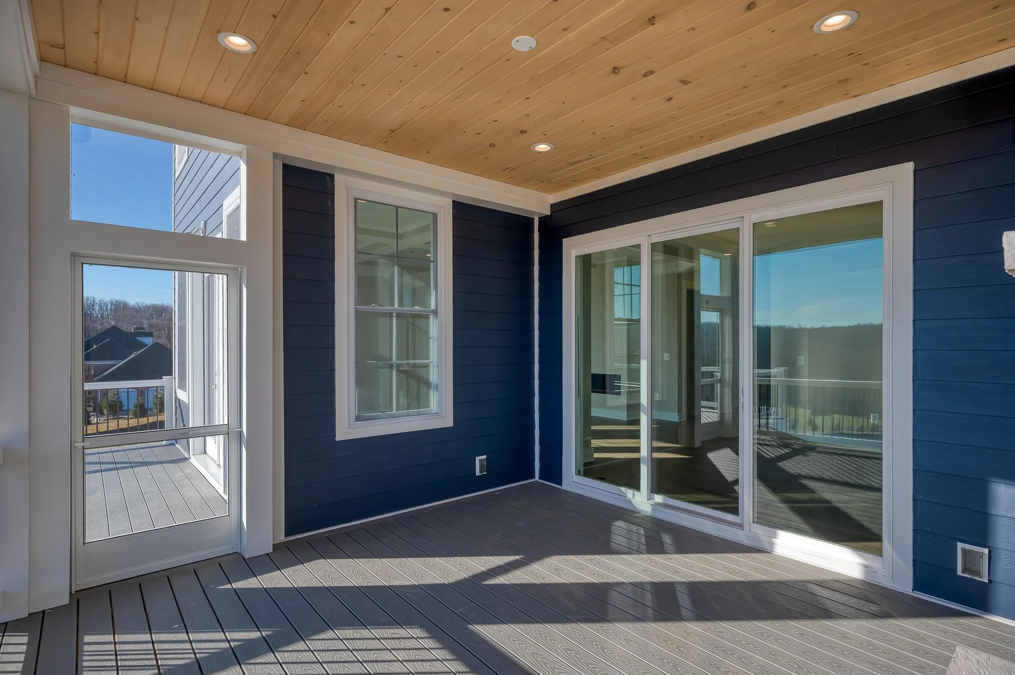 A covered outdoor balcony or porch area with wood flooring, blue siding, white trim, glass sliding doors, a window, and a ceiling with recessed lighting, overlooking neighboring houses and landscape.