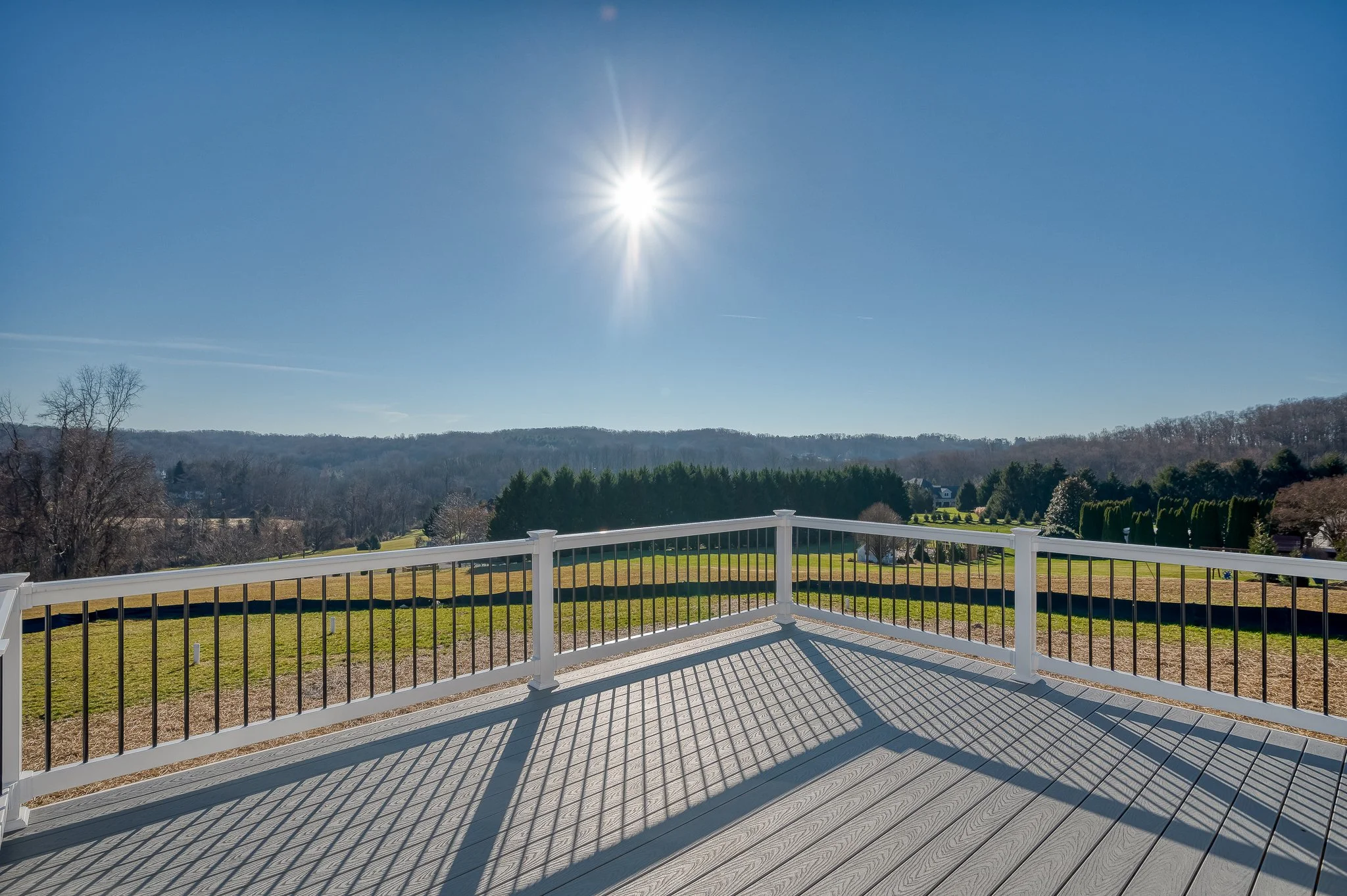 View from a deck with white railing, overlooking a grassy landscape and distant hills under a clear blue sky with bright sun.