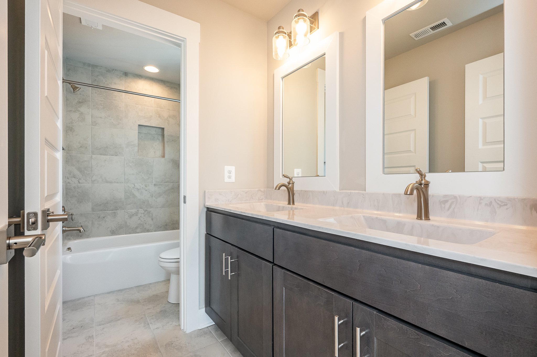 Bathroom with double sink vanity, dark wood cabinets, marble countertop, and two mirrors. Shower with tiled walls and a niche, closed door. Modern lighting fixtures above the mirrors.