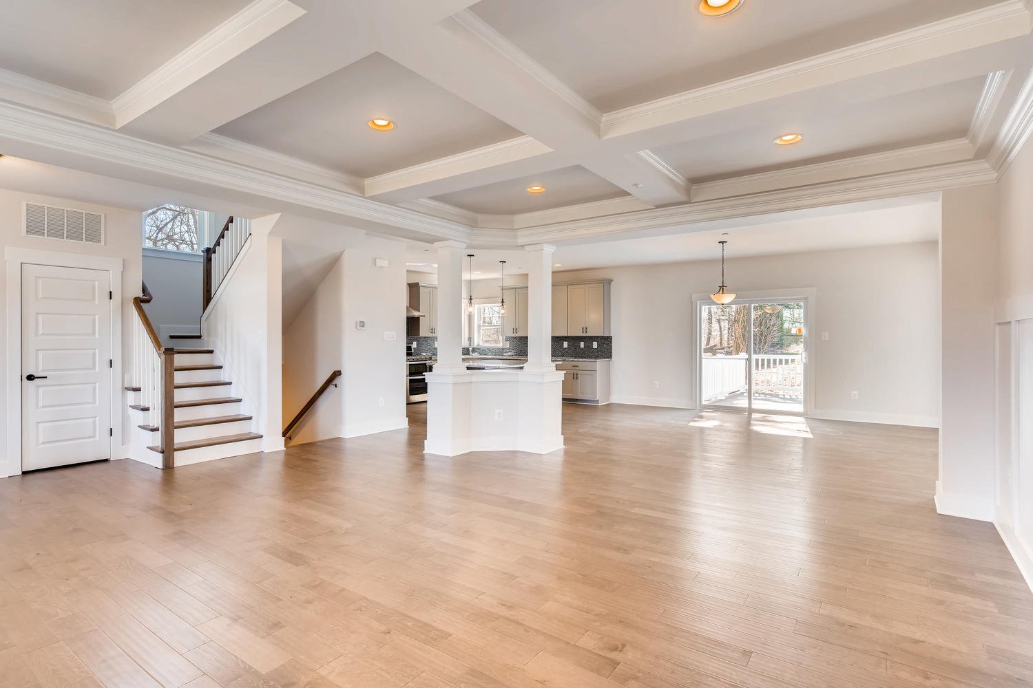 Empty, modern living room and kitchen area with hardwood floors, white walls, recessed lighting, and sliding glass door leading to an outdoor space.
