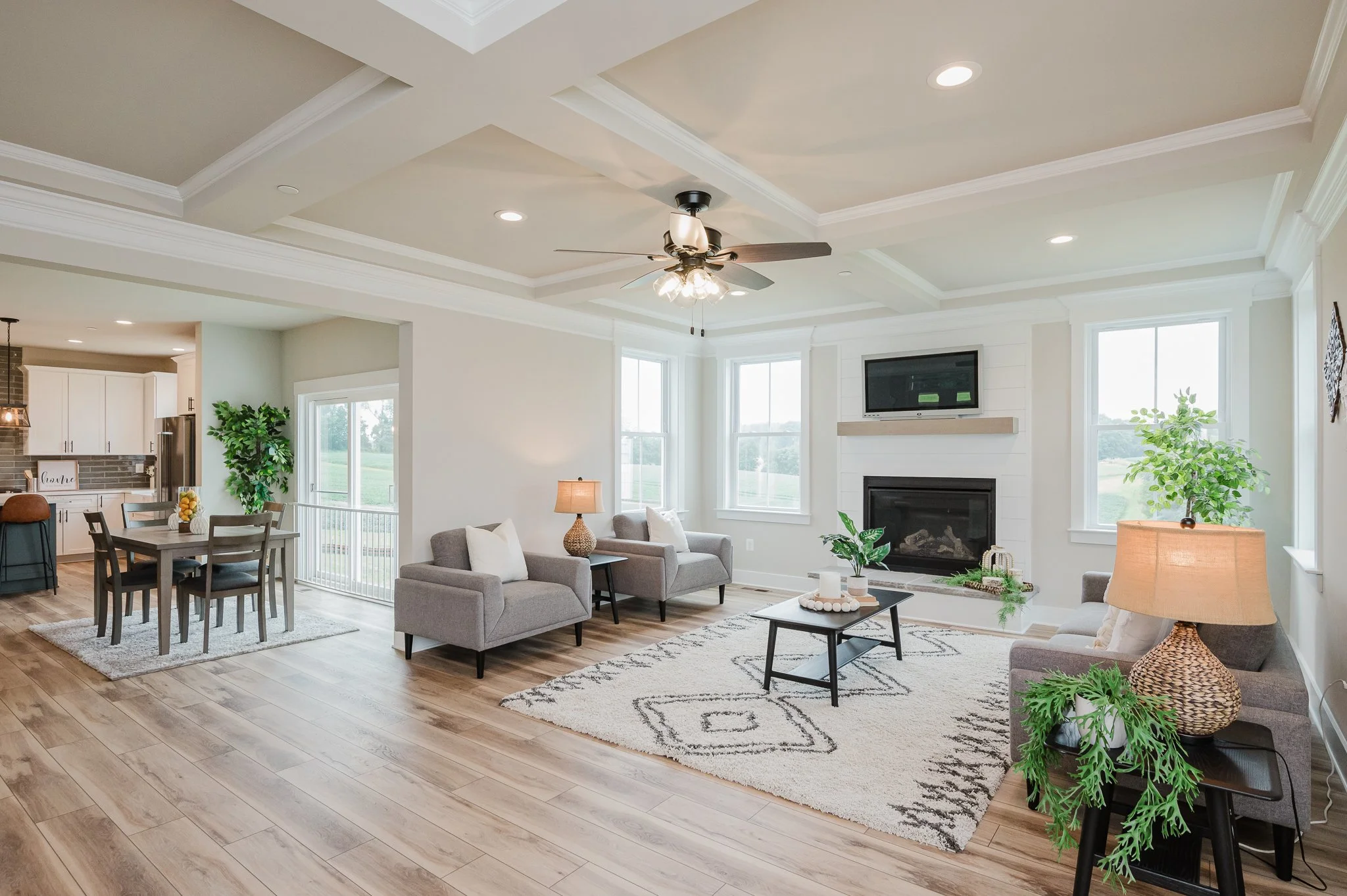 Bright open living room with beige walls and white trim, featuring a fireplace, a ceiling fan, and large windows with a view of the outdoors. The room includes sofas, coffee table, lamps, and decorative plants, with an adjacent dining area and kitche