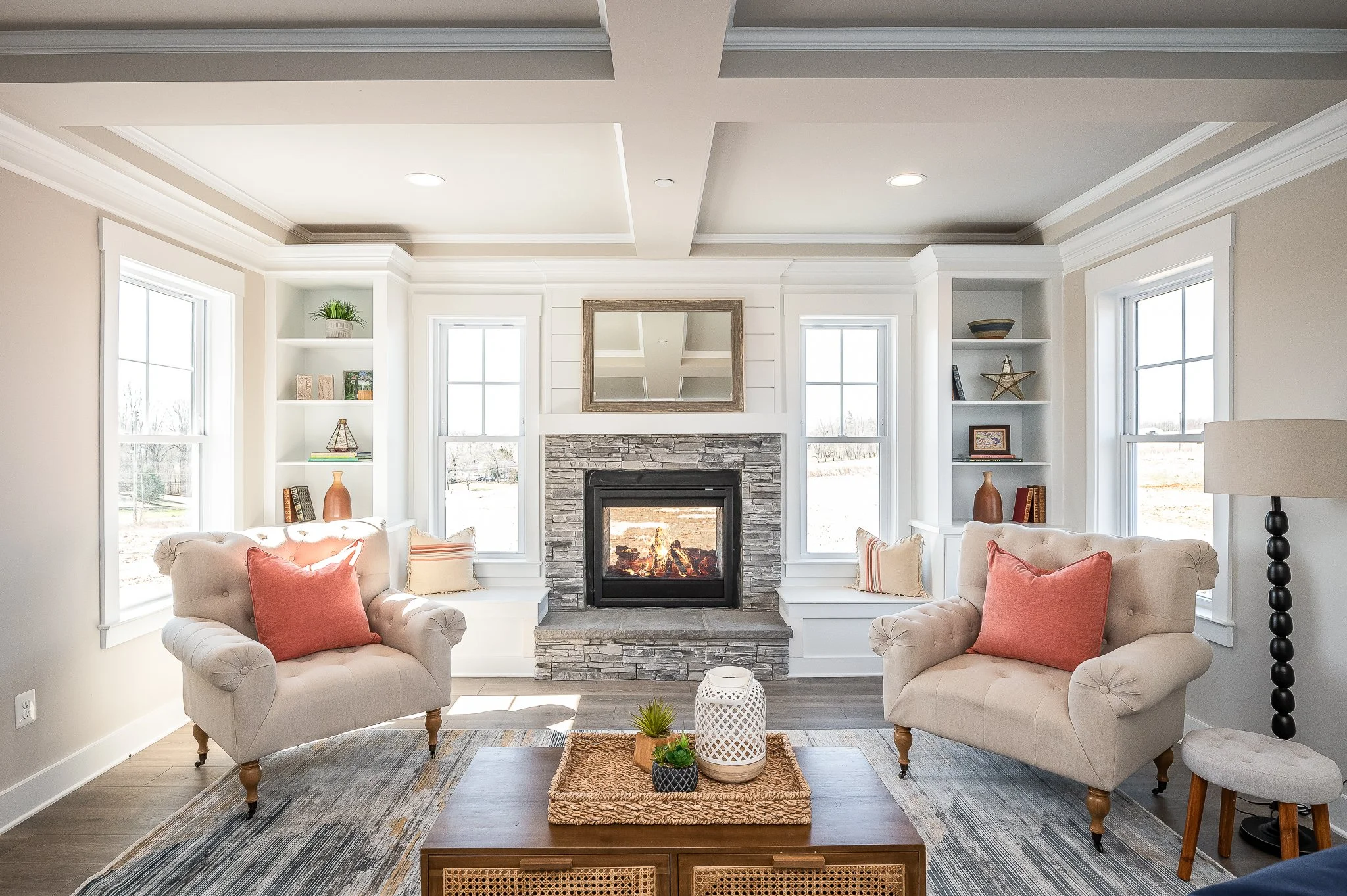 Living room with two beige armchairs with red pillows, a wooden coffee table with decor, a fireplace with a stone surround, and three large windows letting in natural light.
