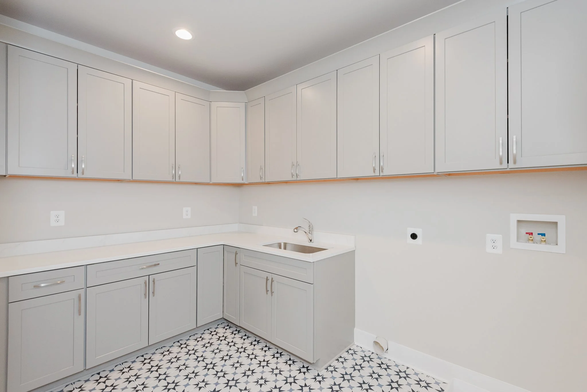 Empty laundry room with light gray cabinets, white countertop, stainless steel sink, and patterned tile floor.