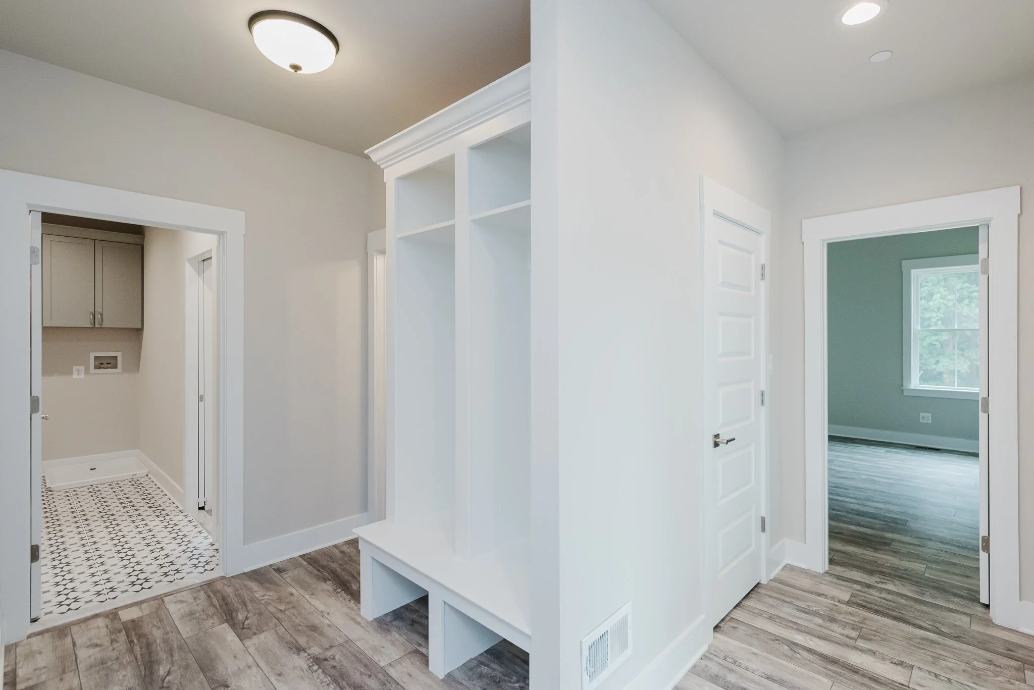 Interior of a modern house with white walls, wood flooring, and a small laundry area with patterned tile floor and gray cabinets.