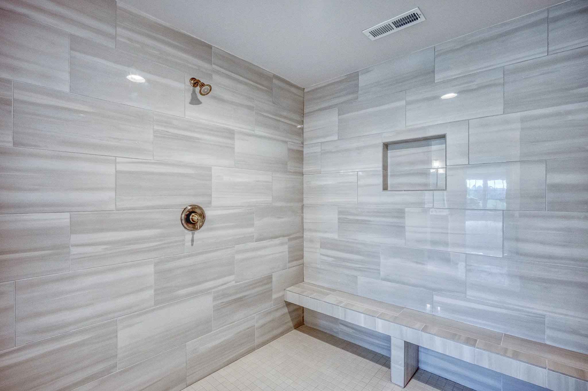 Modern walk-in shower with large light gray tiles on the walls, a built-in niche, a bench, and a ceiling-mounted showerhead.